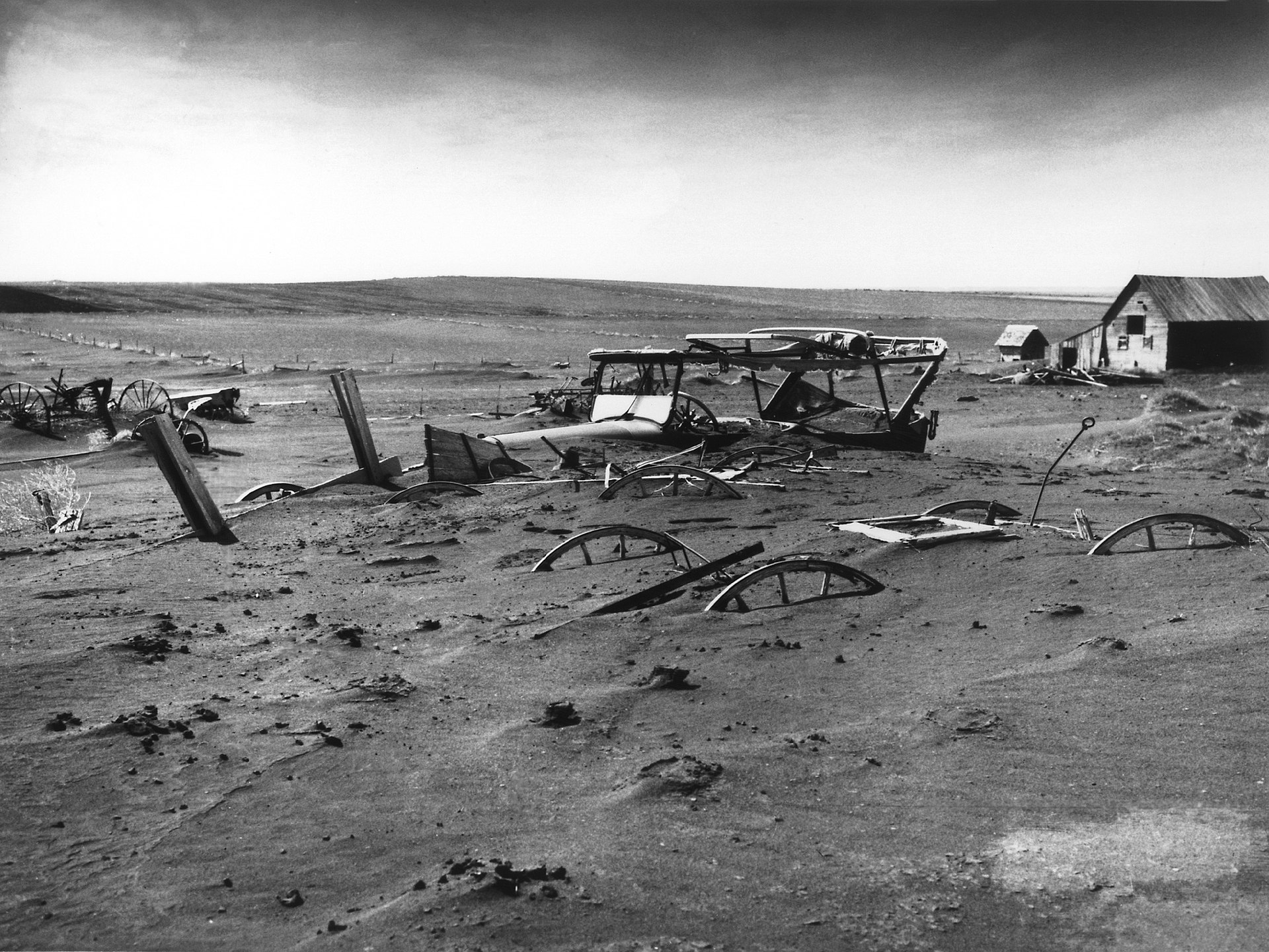 Eine Schwarz-Weiß-Aufnahme verstreuten landwirtschaftlichen Schrotts, darunter Wagenräder, auf einer ariden Ebene mit alten Holzhäusern im Hintergrund unter einem hellen Himmel.