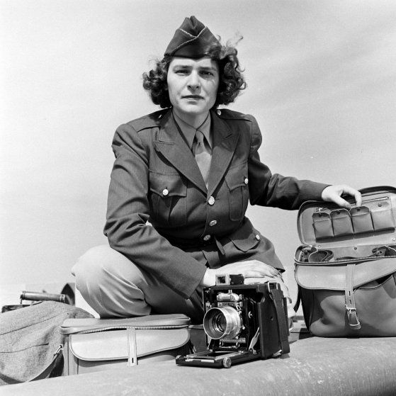 Eine Frau in Uniform sitzt im Freien mit einer Vintage-Kamera vor sich auf einem Tisch und ist von mehreren Gepäckstücken umgeben.