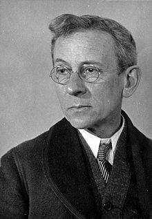 Porträt eines Mannes mittleren Alters mit heller Frisur, der eine Brille und einen dunklen Anzug trägt.