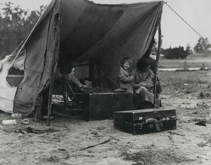 Ein großes Zelt aus Segeltuch steht auf einem offenen Feld und beherbergt mehrere Personen, die in der Nähe gestapelter Holzkisten sitzen.