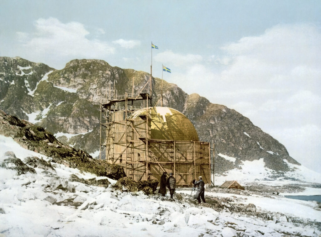 Ein großes, kuppelförmiges Gebäude im Bau steht in einer verschneiten Berglandschaft, wobei mehrere Personen am Fundament arbeiten und Flaggen wehen.