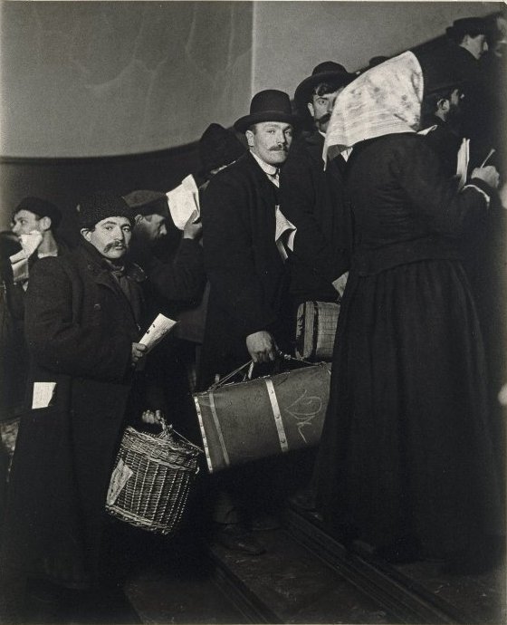 Eine Gruppe von Menschen in historischer Kleidung versammelt sich drinnen, wobei einige Körbe und Kisten tragen.