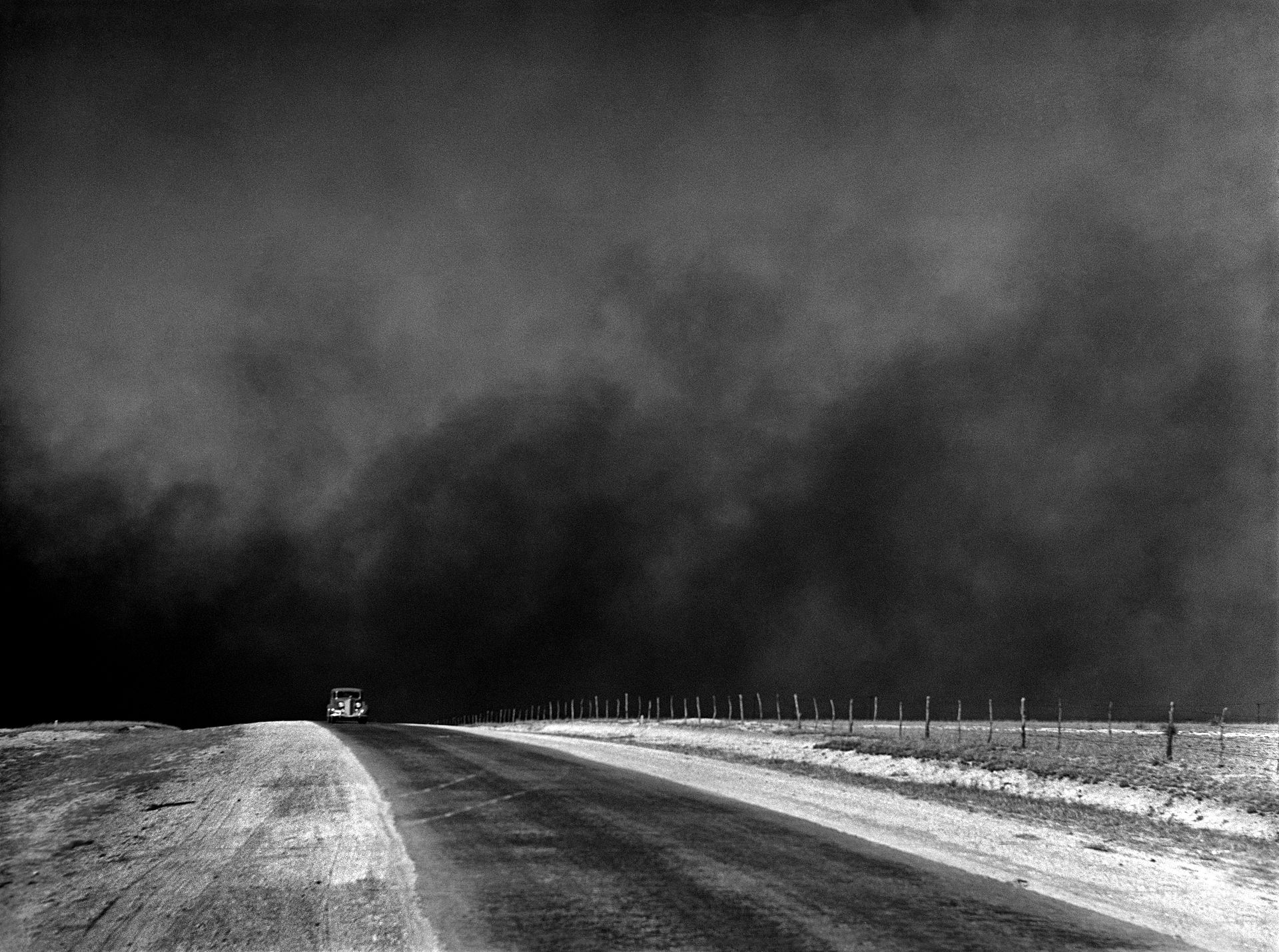 Die Straße erstreckt sich unter einem sehr dunklen Himmel; rechts verläuft ein Zaun und in der Ferne sind zwei Fahrzeuge zu erkennen.