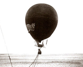 Ein großer, dunkler Ballon schwebt über einer Wasseroberfläche, wobei unten die Befestigungseile und ein Korb zu sehen sind.