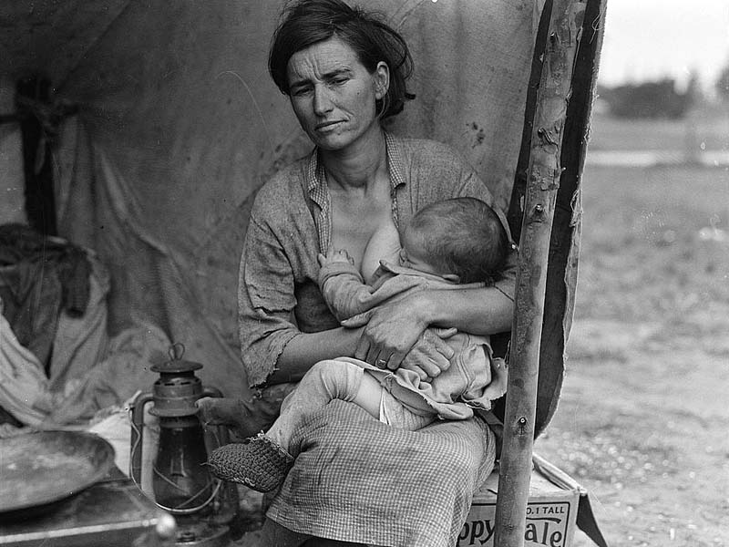 Eine Frau sitzt vor einem Zelt und hält zwei Babys, eines an der Brust und das andere in einem Tuch auf ihrem Schoß.