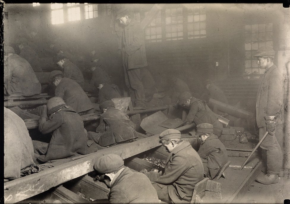 In einer schwach beleuchteten Industriewerkstatt sitzen zahlreiche Arbeiter an langen Tischen und verrichten dort manuelle Tätigkeiten.