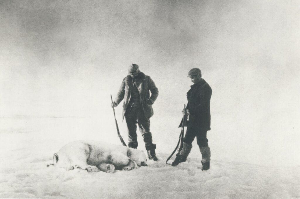 Zwei Männer in Winterkleidung stehen auf Schnee und blicken zu einem liegend ruhenden Hund.