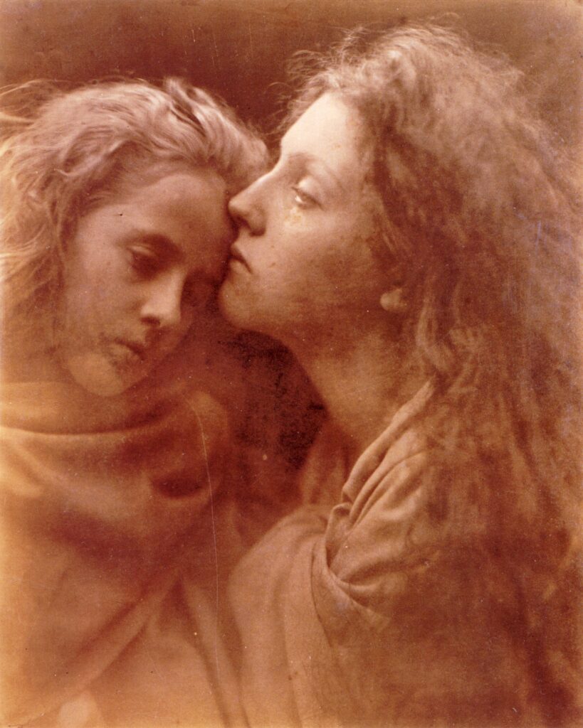 Zwei Frauen mit langem Haar sind in einem Sepia-Tonporträt eng zusammengehalten, wobei die Frau rechts leicht aufblickt und die andere Frau sich zu ihr neigt.