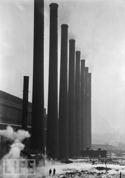 Eine lange Reihe industrieller Schornsteine steht unter einem bewölkten Himmel und verjüngt sich nach hinten, wobei kleine Figuren am basischen Rauch sichtbar sind.