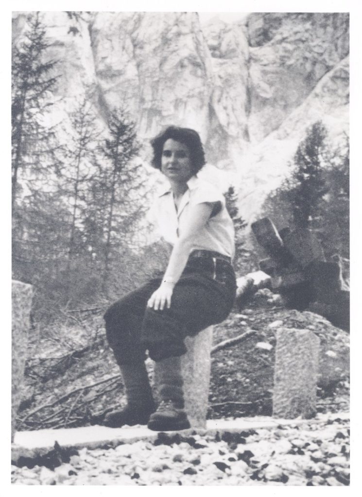 Eine Schwarz-Weiß-Fotografie zeigt eine Frau, die draußen auf einem Stein sitzt und vor einer Kulisse aus Wald und Felswänden posiert.