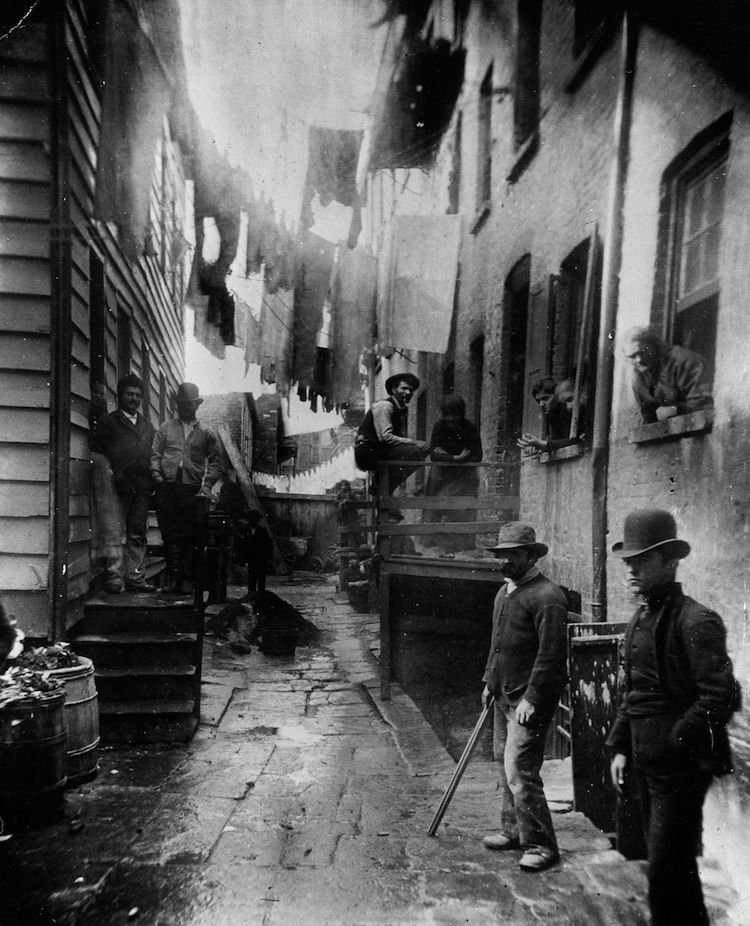 Eine enge Gasse mit Gebäuden auf beiden Seiten, an der Wäsche zwischen den Häusern hängt und mehrere Personen zu sehen sind.