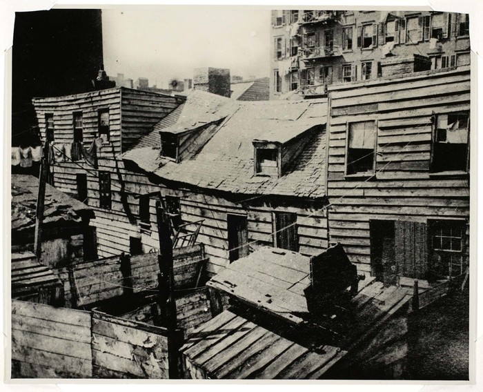 Ein Schwarz-Weiß-Foto von dicht gedrängten, alten Holzhäusern mit Satteldächern vor dem Hintergrund moderner Hochhäuser.