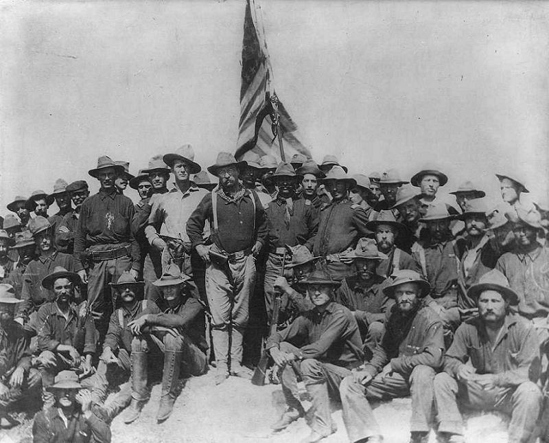 Eine große Gruppe von Männern in Uniform sitzt und steht im Freien vor einer großen Flagge.