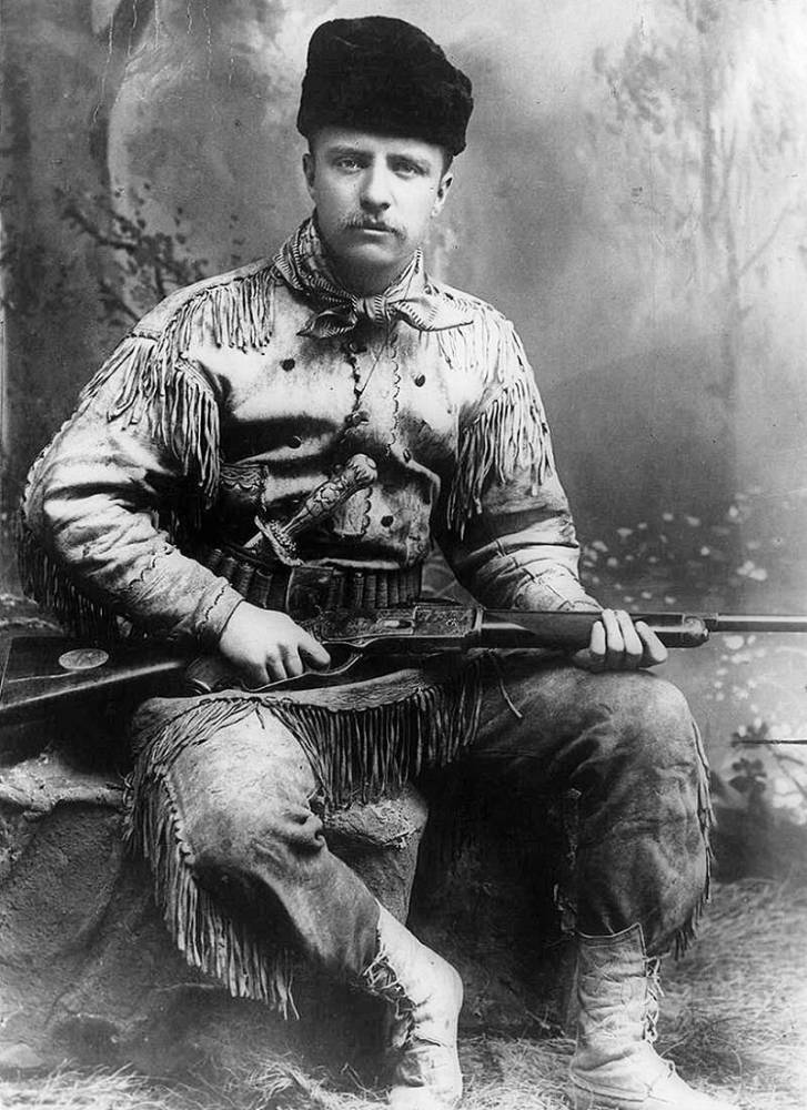 Ein Mann in fransiger Kleidung und einem dunklen Hut sitzt mit einem Gewehr vor einem gemalten Waldhintergrund.